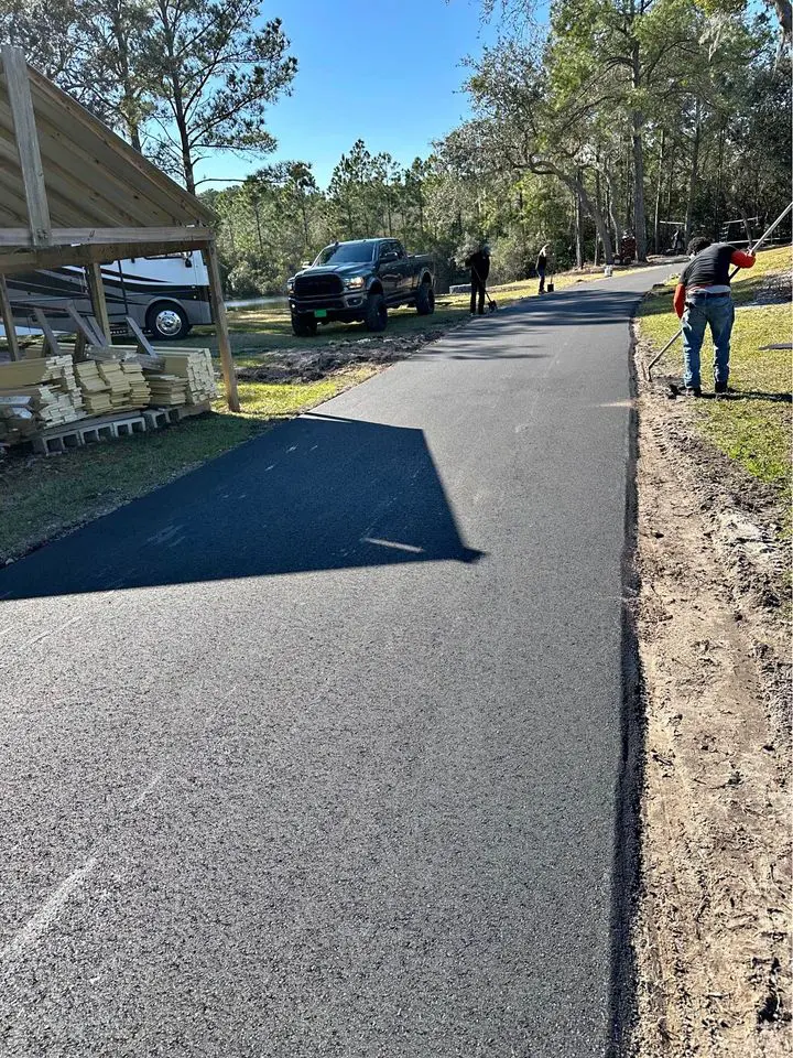 Workers from a licensed asphalt contractor are paving an outdoor path with asphalt in a wooded area. A black truck is parked nearby, while some workers stand and others work on the new surface. Stacks of wooden planks are stored under a wooden shelter to the left. Trees are visible in the background.
