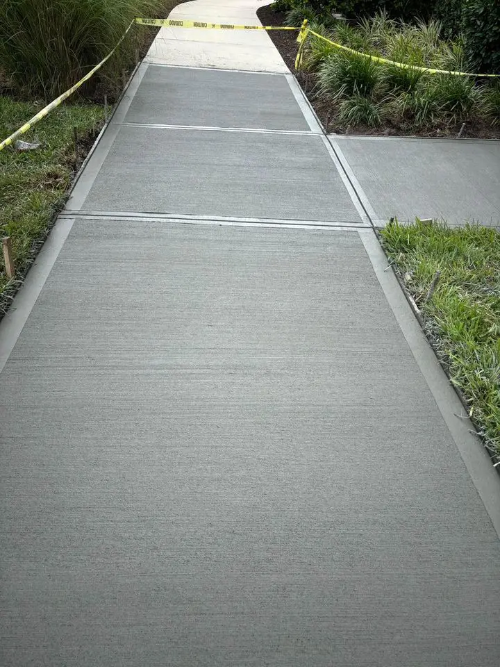 Freshly laid and curing concrete sidewalk enclosed with yellow caution tape on a grassy path in Delray Beach, expertly managed by a licensed asphalt contractor.