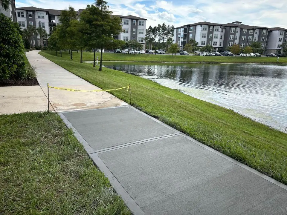 A newly paved concrete sidewalk section blocked by caution tape beside a grassy area and a pond, with an apartment complex in the background. The sky is partly cloudy, reflecting the expertise of licensed asphalt contractors in Deerfield Beach.