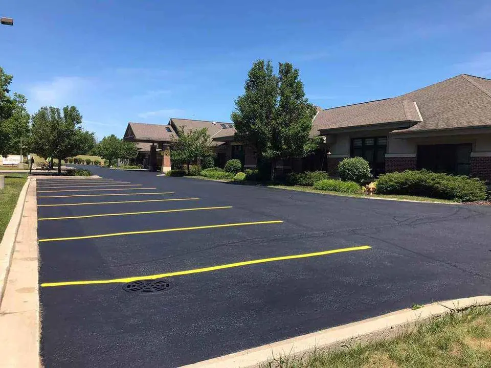 A freshly paved parking lot, courtesy of licensed asphalt contractors, features clearly marked yellow lines in front of a row of single-story office buildings with brick facades and gabled roofs in Deerfield Beach. The area is landscaped with green bushes and trees under a clear blue sky.