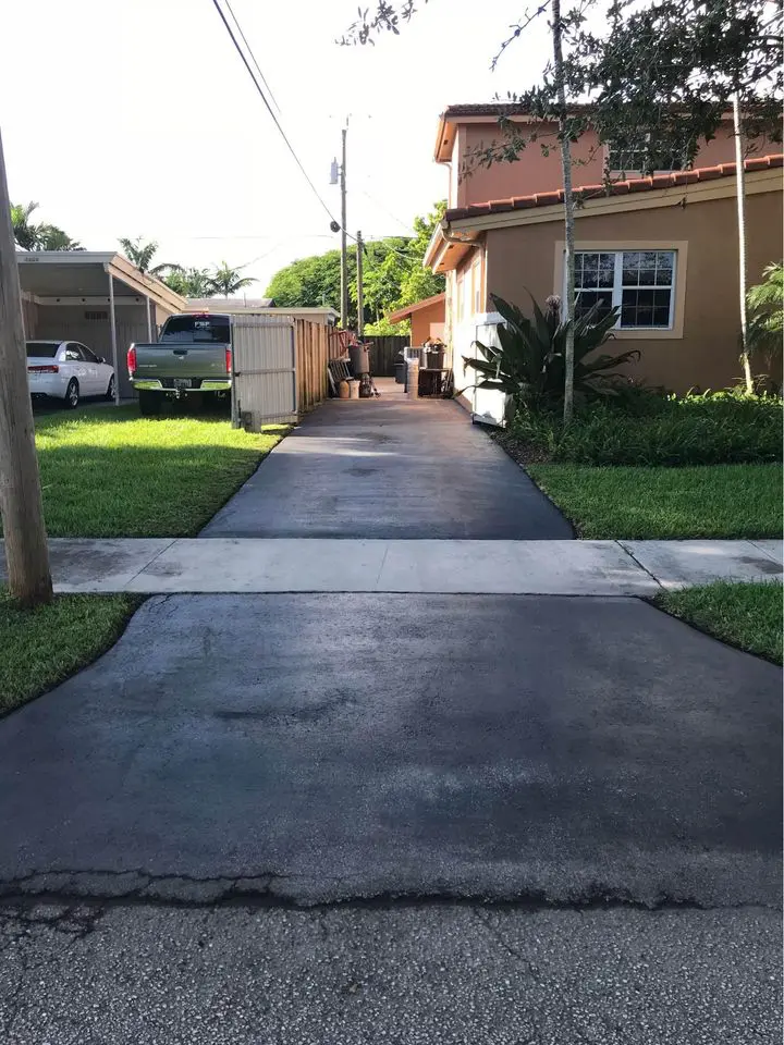 A driveway leading to a beige house with a tiled roof showcases the work of asphalt contractors from Deerfield Beach. A gray truck is parked to the left of the house, and a fence runs alongside the newly paved driveway. Potted plants and trees surround the area, as a person walks towards the house.