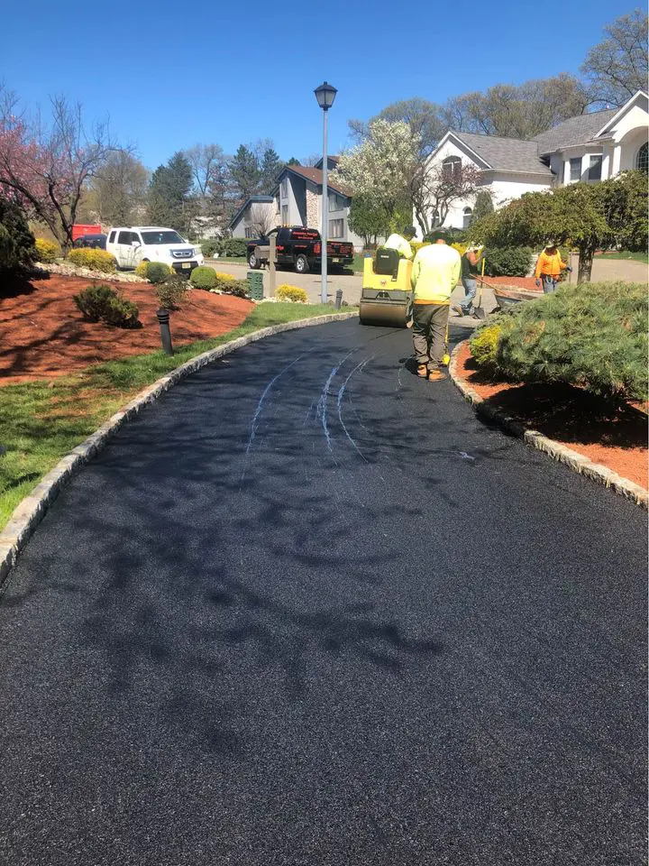 Licensed asphalt contractors from Deerfield Beach are laying fresh asphalt on a residential driveway. The driveway is bordered by landscaped gardens with shrubs and a lamp post. A roller machine is being used by one of the workers to smooth the new surface. Other workers and a few vehicles are in the background.